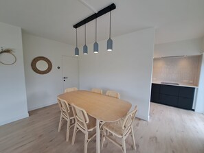 Ein Essbereich mit Holztisch und Korbstühlen, einer weißen Tür sowie einer Küche mit dunklen Schränken.