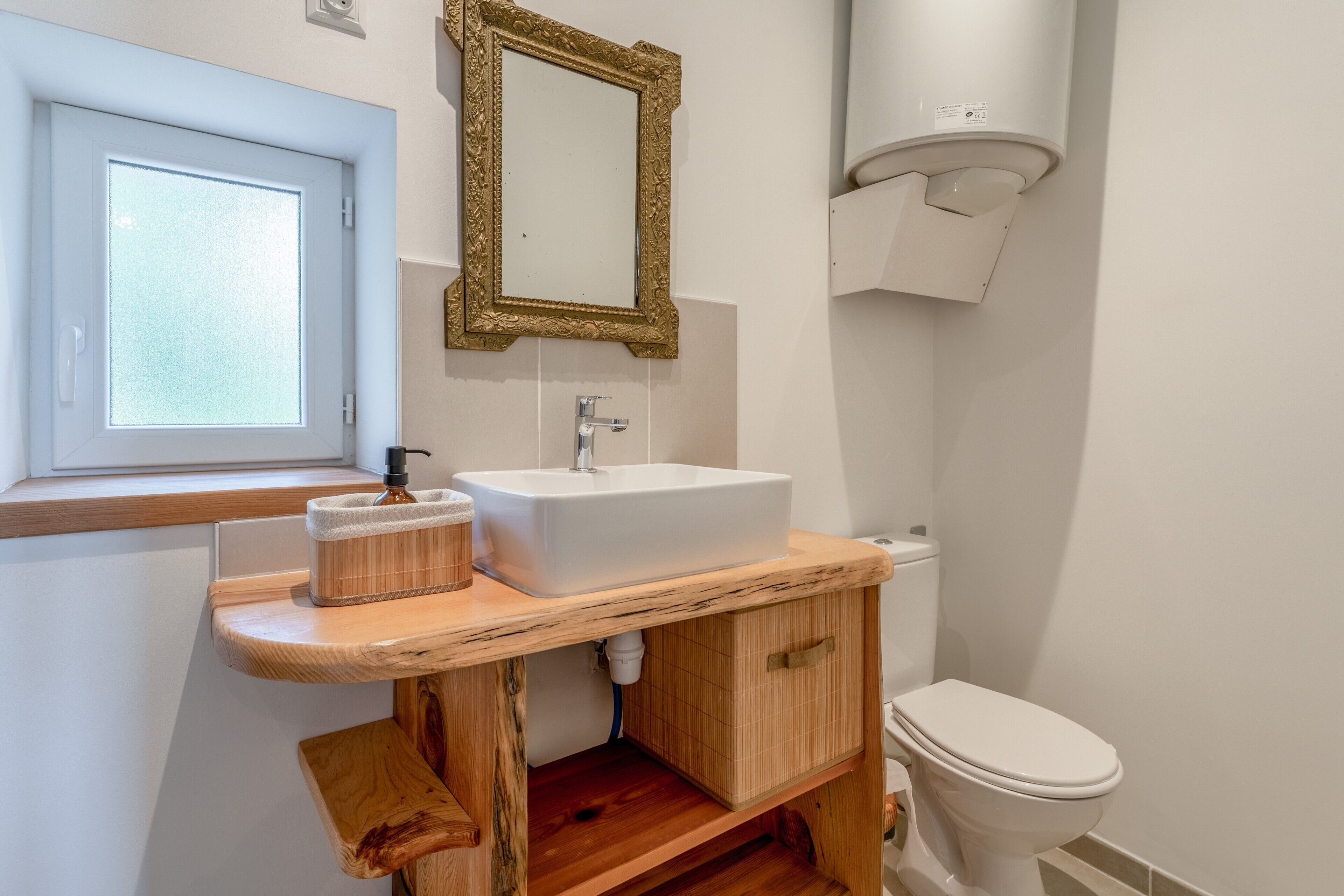 Een badkamer met een witte wastafel, houten opbergmeubel, een spiegel, toilet en een raam met geëtste glas.
