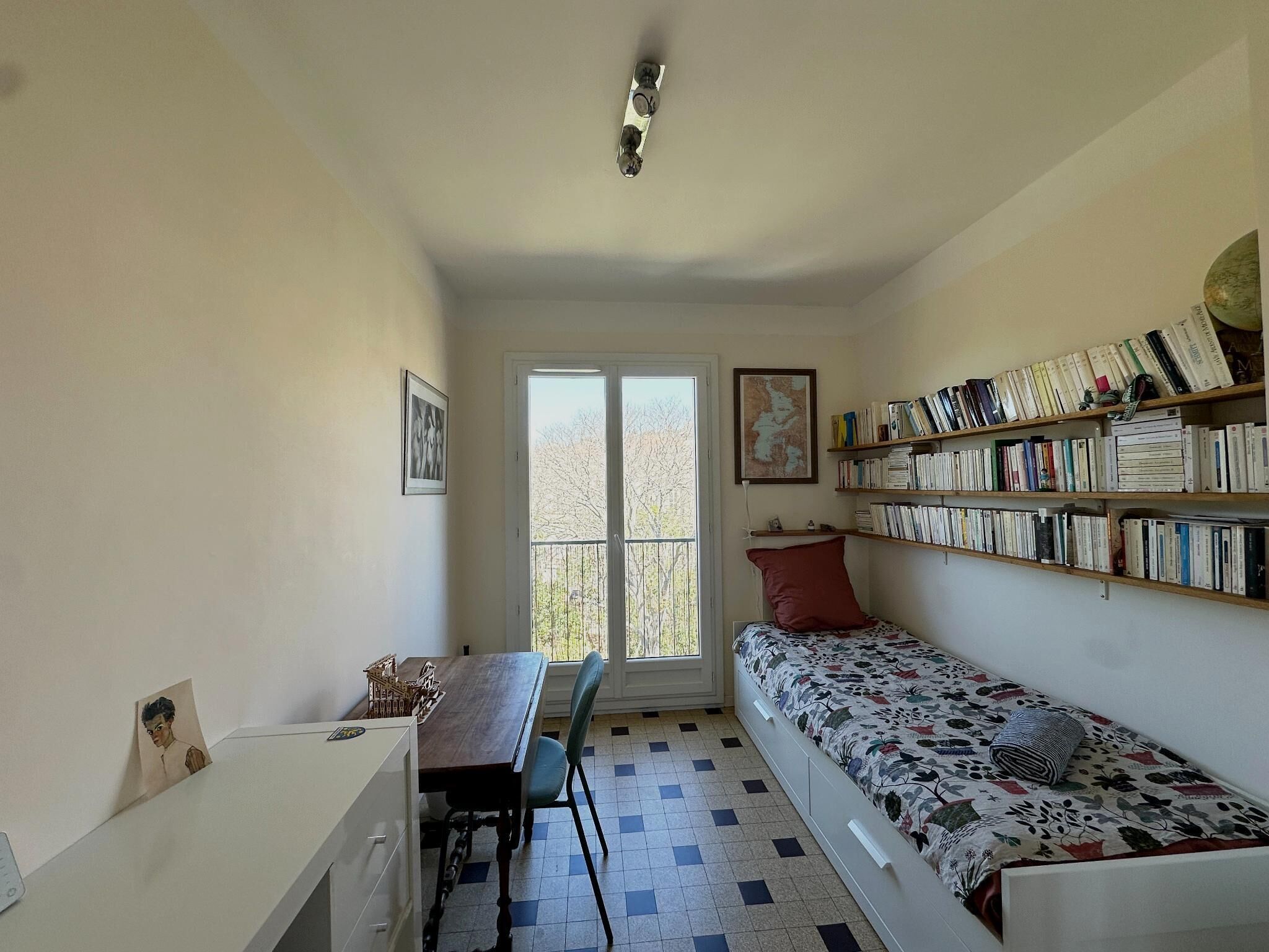 Una habitación pequeña con una cama, un escritorio, una silla, una estantería y una ventana.