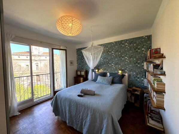 Un dormitorio con una cama grande, una ventana con vistas a edificios, una estantería y una lámpara colgante.