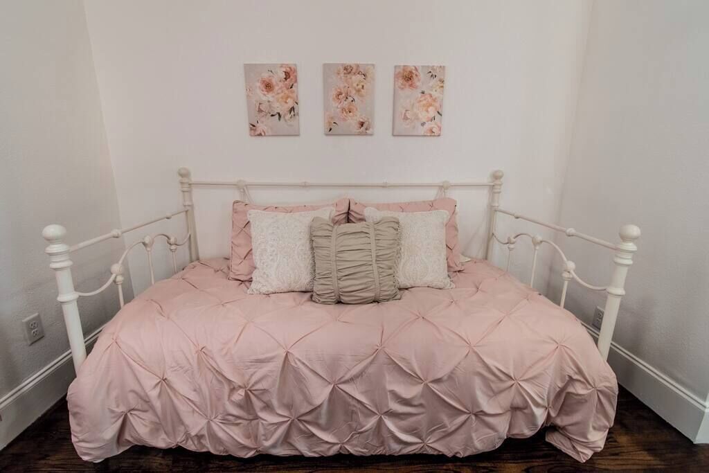 A bed with a pink quilt and white pillows, a white metal frame, and three floral paintings on the wall.