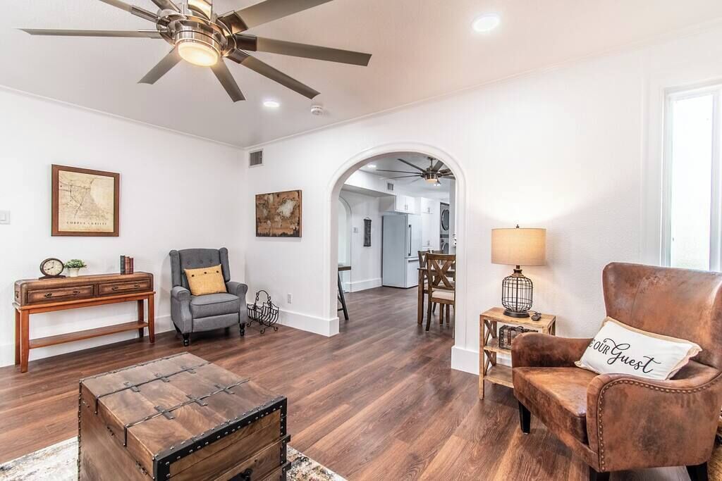 Un vaste salon doté d’un plancher en bois, d’un ventilateur de plafond, d’une table basse en bois, d’un fauteuil gris et d’un fauteuil en cuir marron, ainsi qu’un coin repas visible à travers une arche.