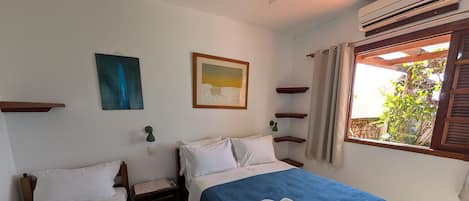 A bedroom with two beds, a window with curtains, a wall-mounted air conditioner, and a small table with a lamp.