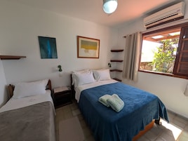 A bedroom with two beds, a window with curtains, a wall-mounted air conditioner, and a small table with a lamp.