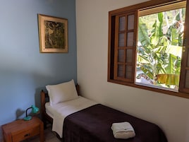 A single bed with a dark bedspread, a white pillow, a small bedside table with a lamp, a window with a view of banana trees, and a framed picture on the wall.