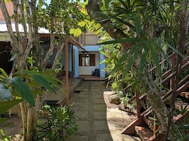 Un sendero de piedra conduce a una casa de color azul, rodeada de abundante vegetación y una escalera de madera.