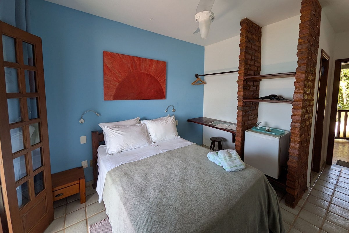 Un dormitorio con cama, un escritorio, una lámpara colgante, un cuadro abstracto rojo y un pilar de ladrillo.