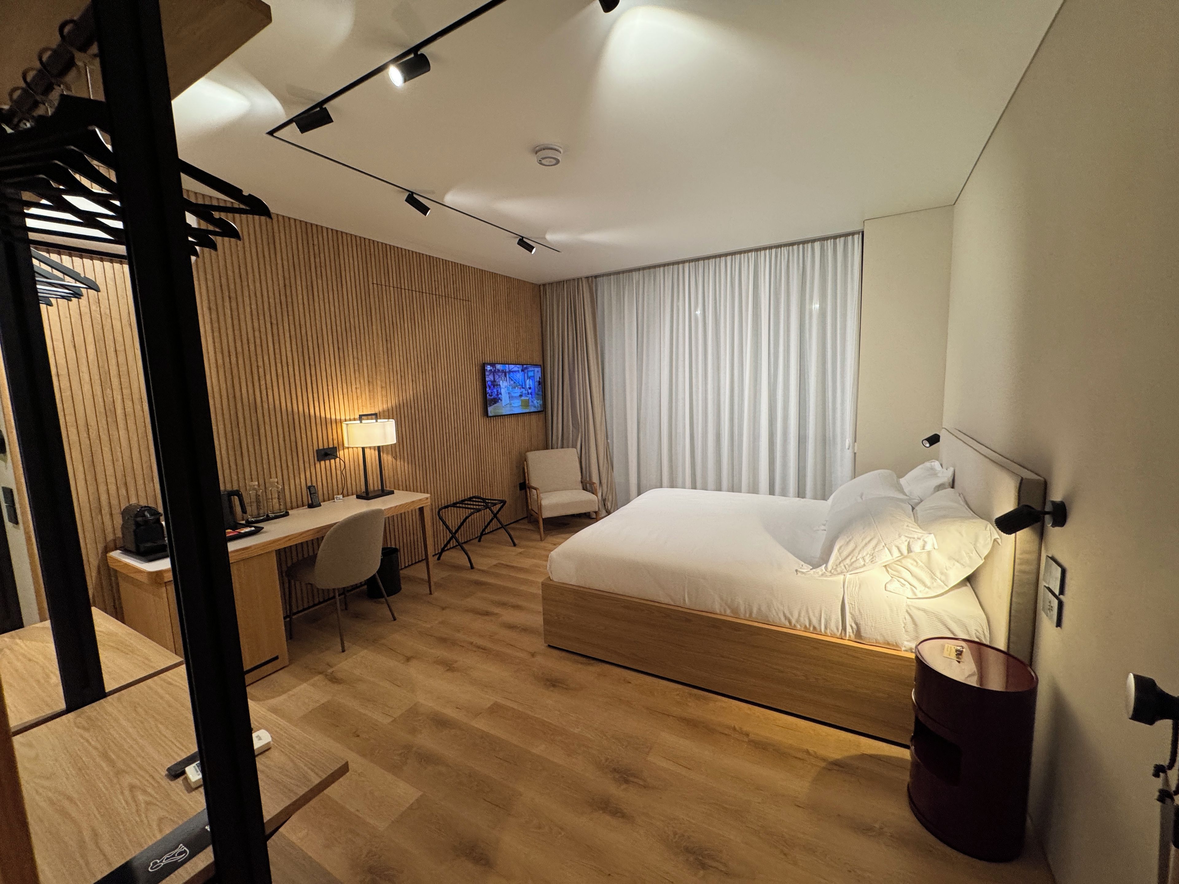 A modern hotel room with a wooden floor, a bed with white linens, a desk with a lamp, a chair, a television on the wall, and a window with curtains.