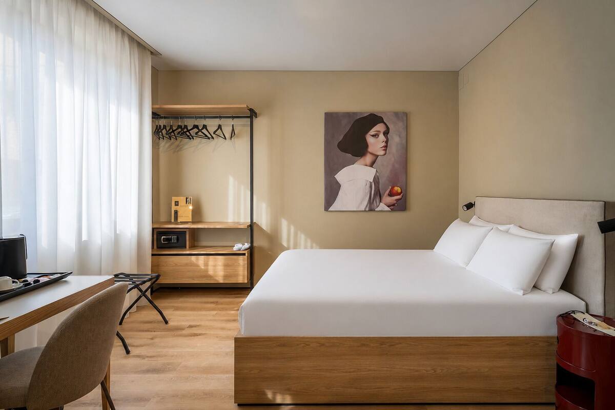 Habitación de hotel con cama, escritorio, silla, perchero y un cuadro en la pared.