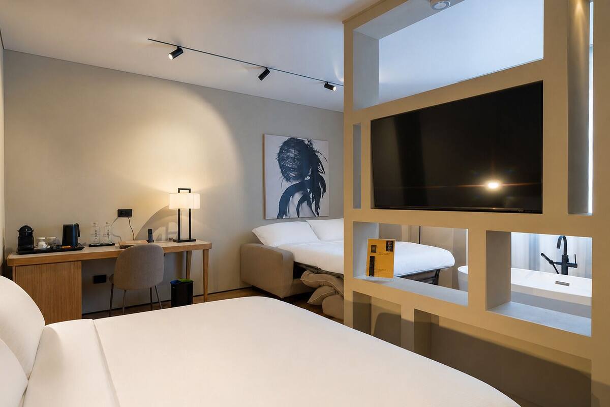 Una habitación de hotel moderna con cama, escritorio, silla, lámpara, televisor y espejo de baño.