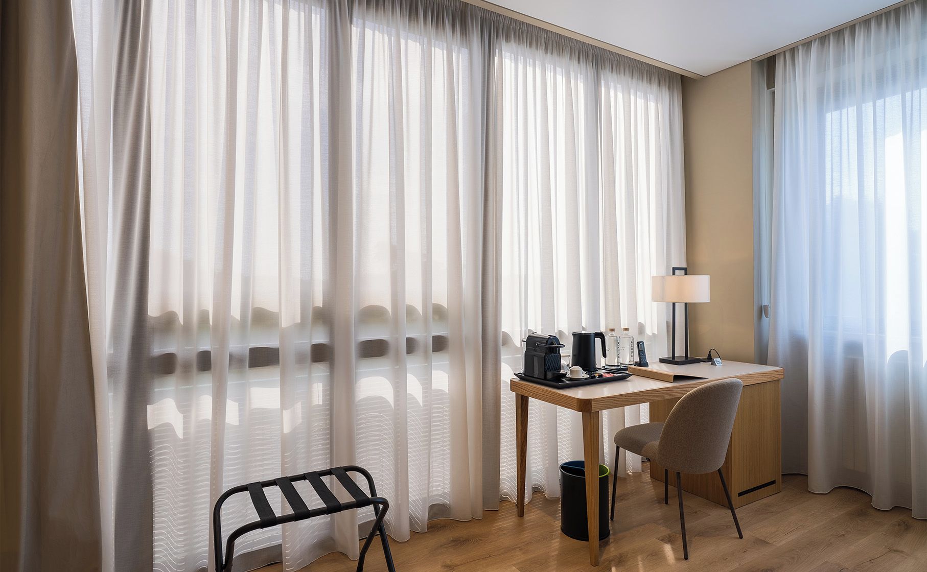 Habitación de hotel con un escritorio de madera, una silla, una lámpara, una cafetera y un cubo de basura.