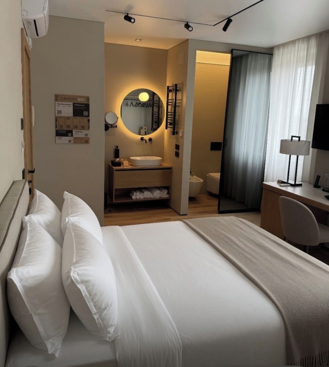 A hotel room with a bed, a desk, a chair, a bathroom, and a large mirror.