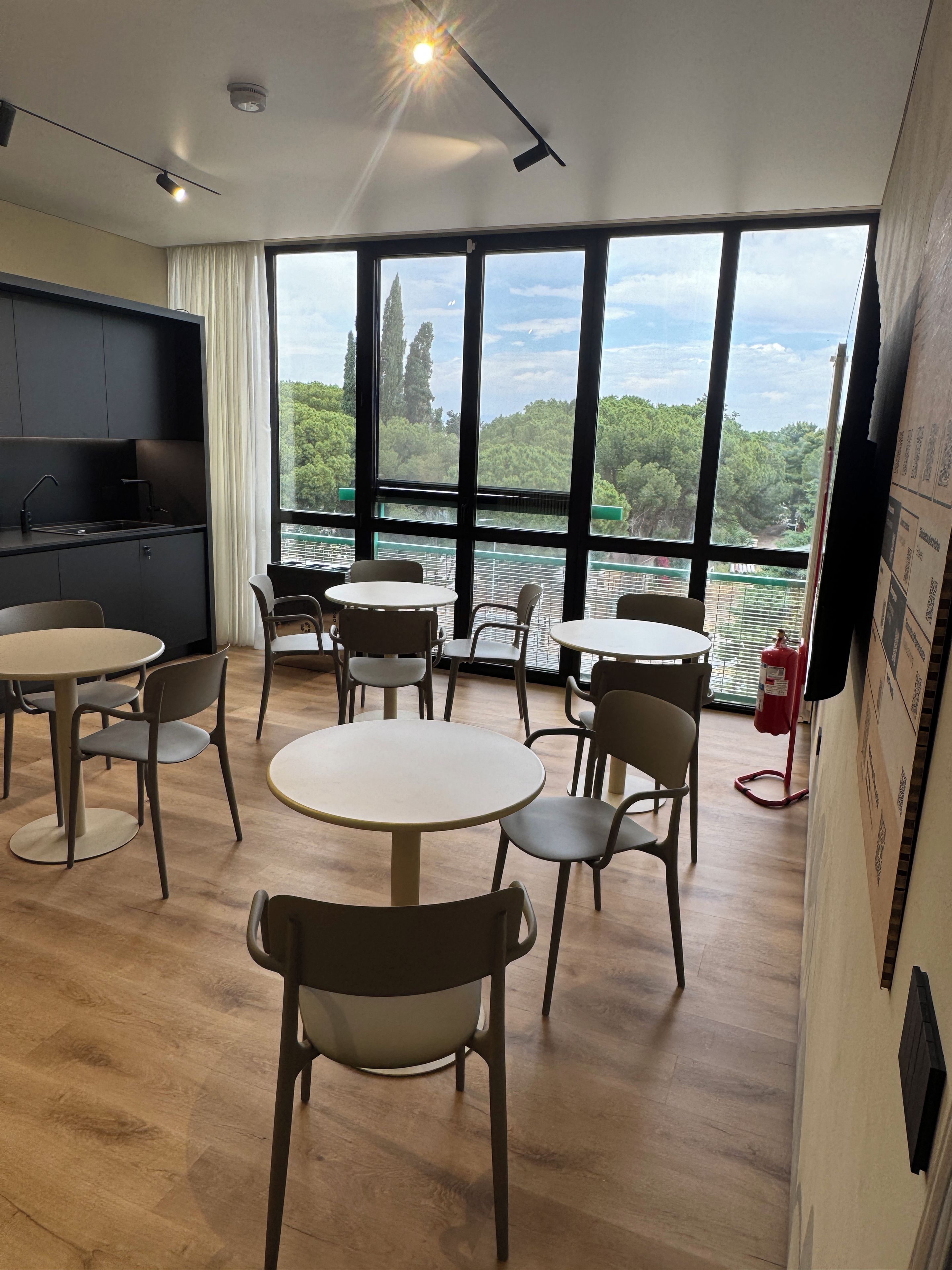 A modern room with a kitchen area, multiple round tables, and chairs, and large windows offering a view of trees and a distant landscape.