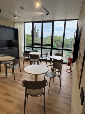 A modern room with a kitchen area, multiple round tables, and chairs, and large windows offering a view of trees and a distant landscape.
