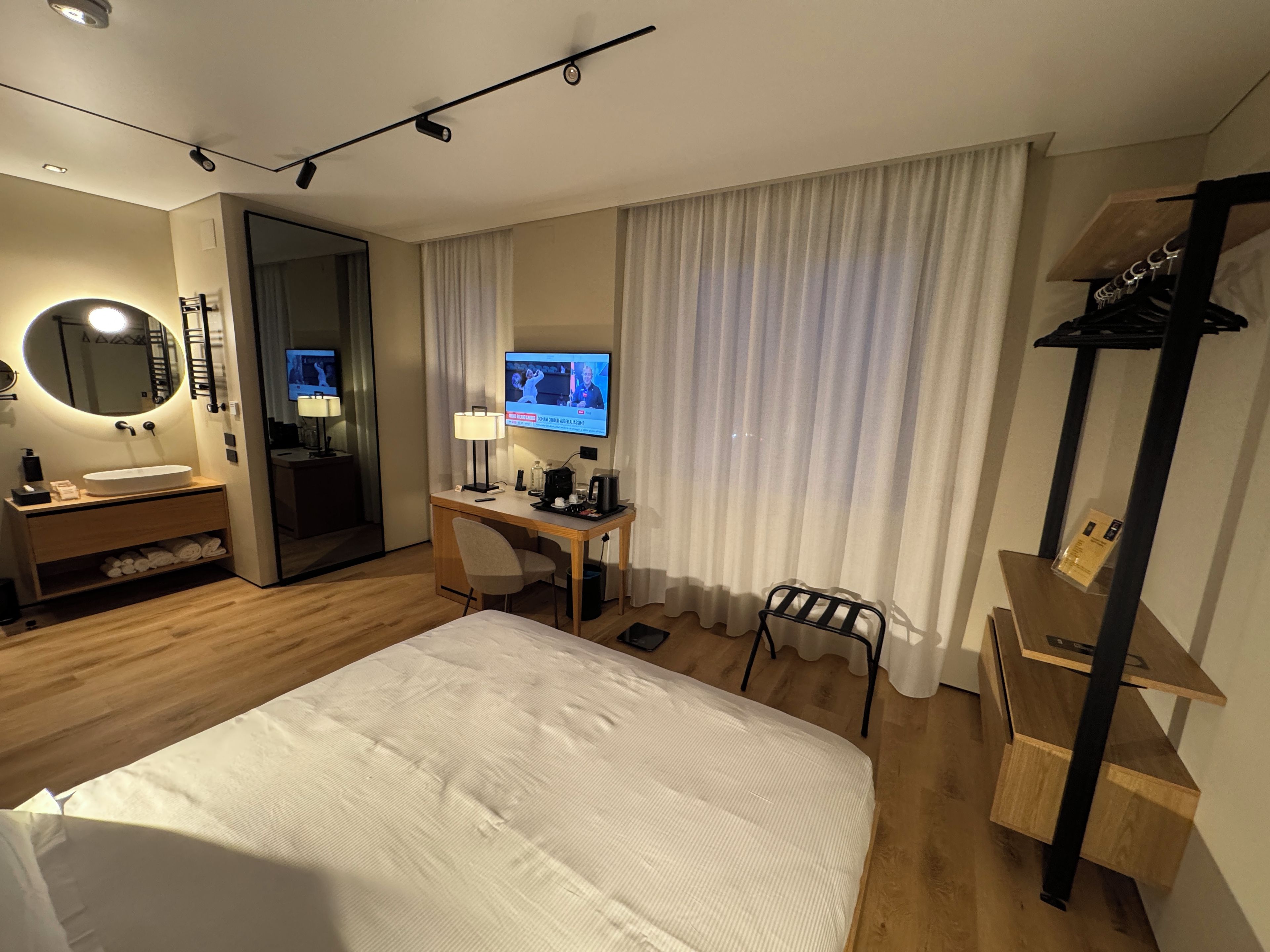 Habitación de hotel con cama, escritorio con computadora, televisor, baño con lavamanos y espejo, y perchero.