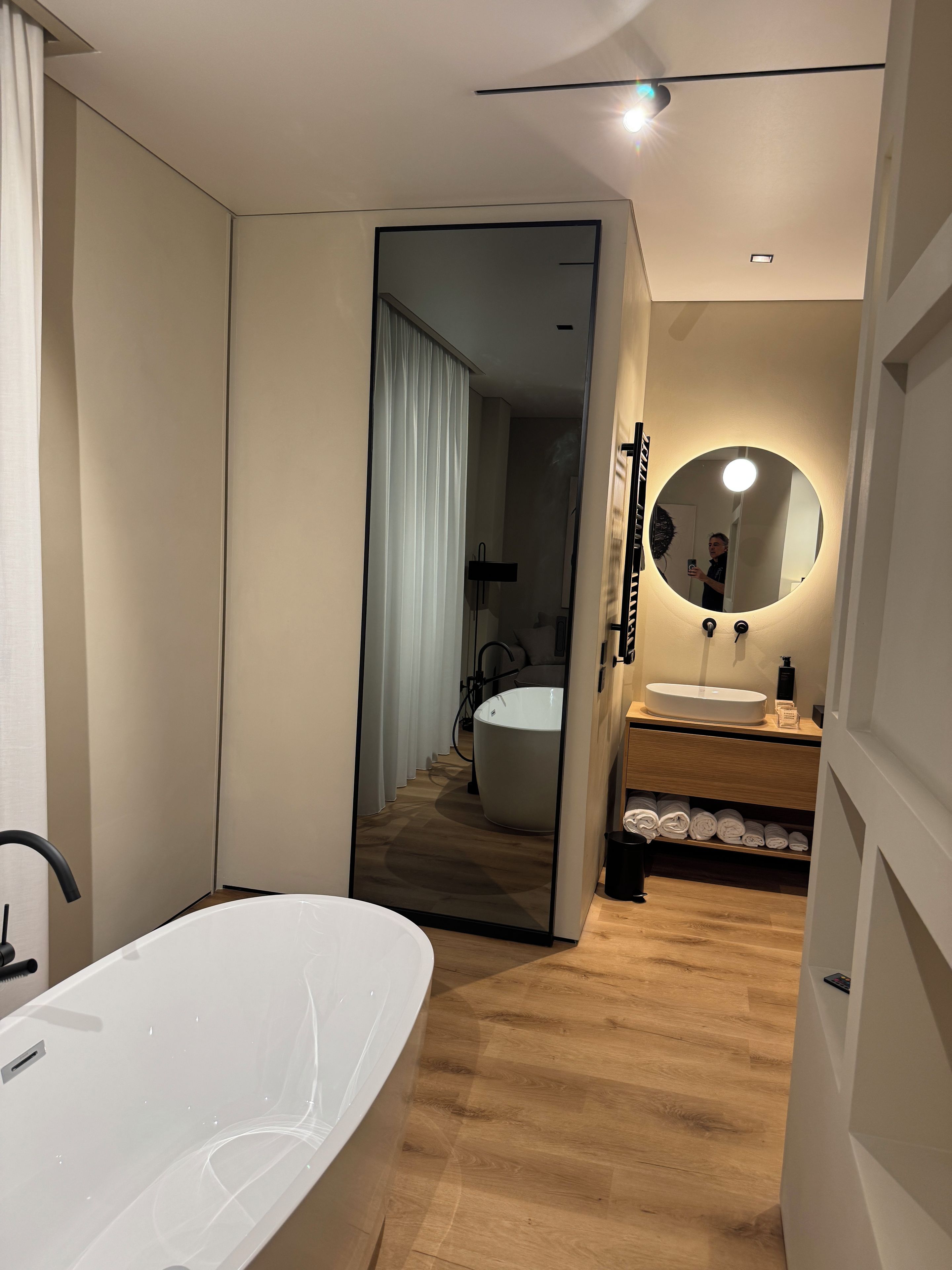 A modern bathroom with a large bathtub, a round mirror, and a wooden vanity.