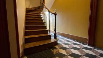 Un escalier avec des marches en bois et une balustrade noire, menant à un palier dont le sol est recouvert d’un motif à carreaux.