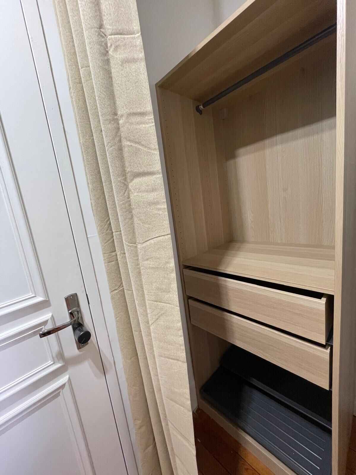 A room with a white door, beige curtains, and a wooden wardrobe with three drawers and a hanging rod.