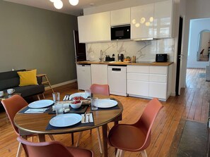 Een moderne keuken met witte kasten, een magnetron en een vaatwasser. Er is een eethoek met een houten tafel en stoelen.