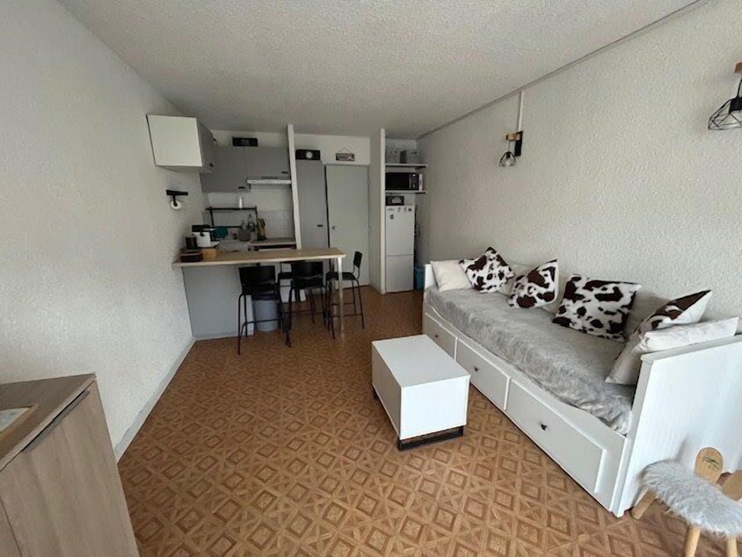 Een ruimtelijk ingerichte leefruimte met een kitchenette, een bank voorzien van decoratieve kussens, een eethoek en een vloerkleed met een patroon.