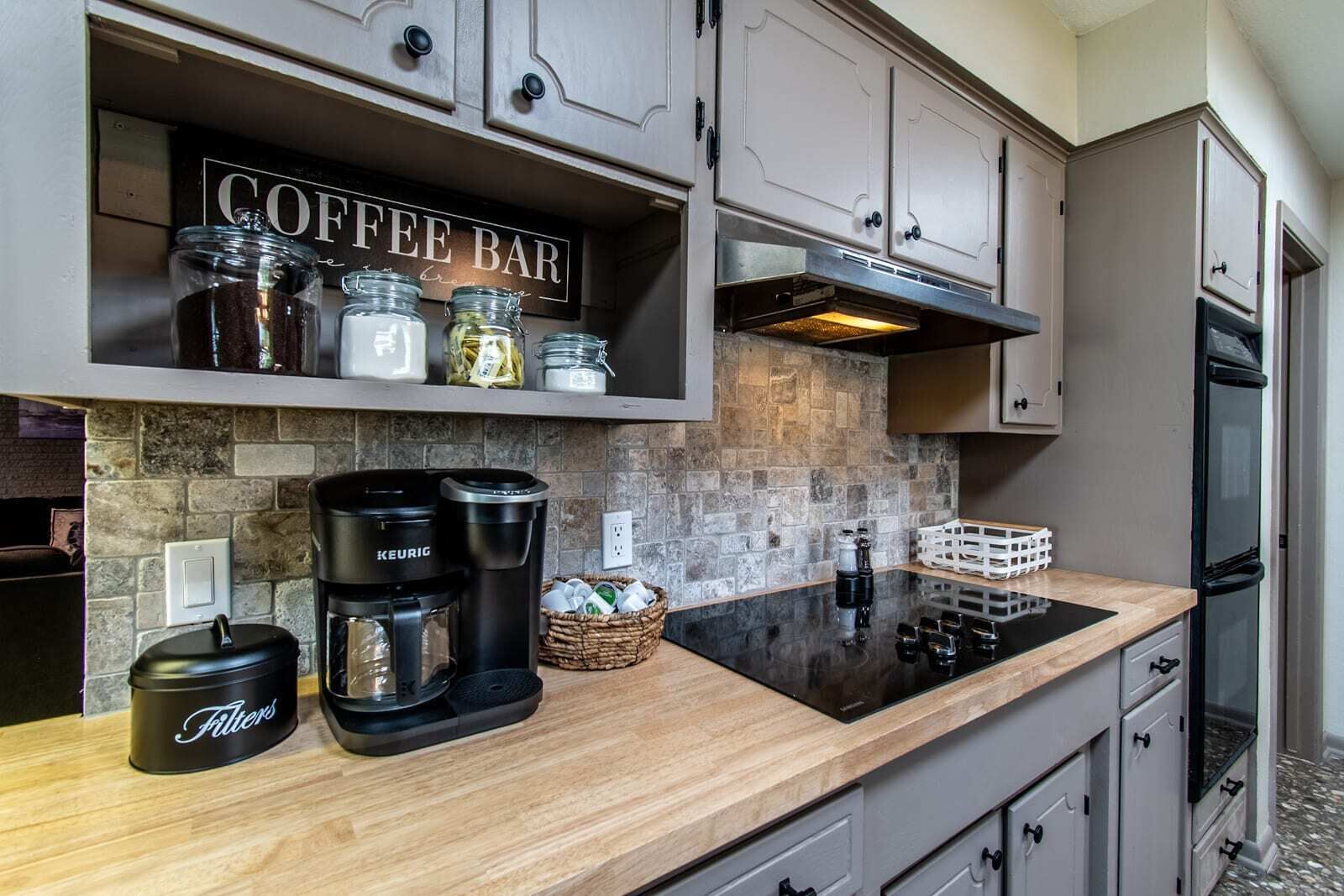 A kitchen with a coffee bar, a Keurig coffee maker, a filter container, and a stone backsplash.
