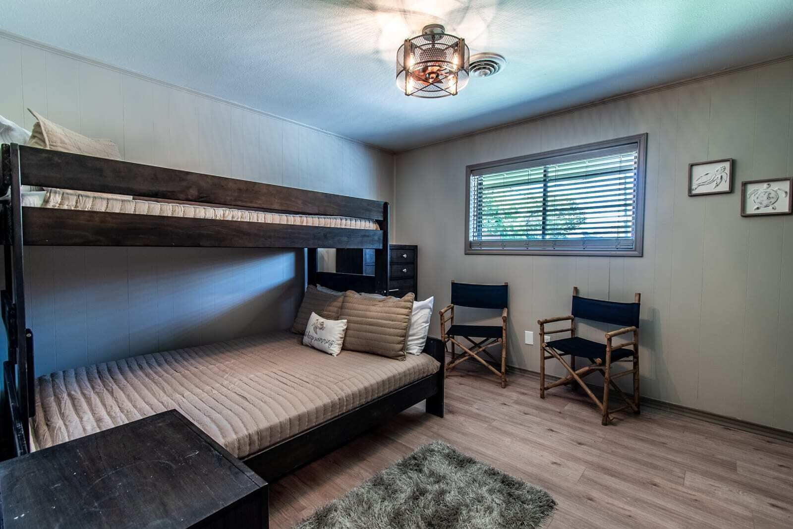 A bunk bed room with a desk, two chairs, a window with blinds, and a ceiling light fixture.