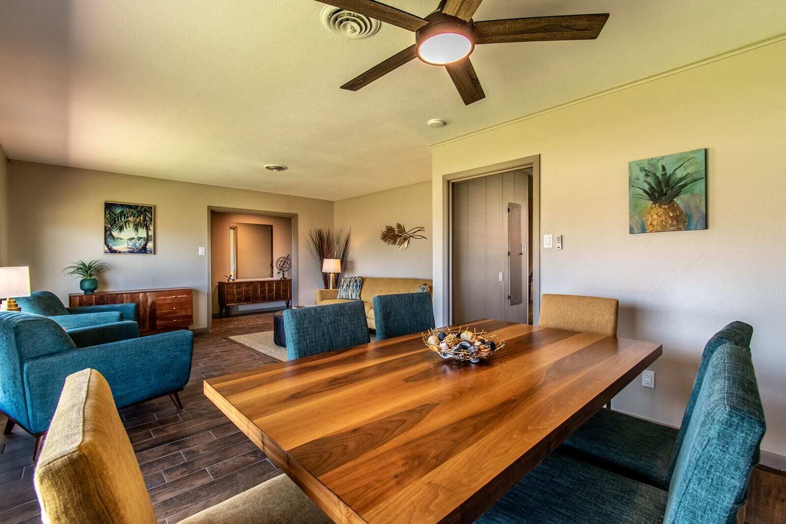 A dining area with a wooden table, blue upholstered chairs, a ceiling fan, and artwork on the walls.