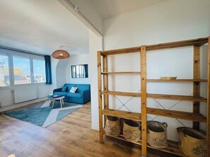 Ein Zimmer mit einem blauen Sofa, einem Holzregal und Blick auf Gebäude durch große Fenster.