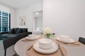 Un espace de repas moderne avec une table ronde dressée pour quatre personnes, un canapé en arrière-plan et une vue sur le paysage urbain à travers de grandes fenêtres.