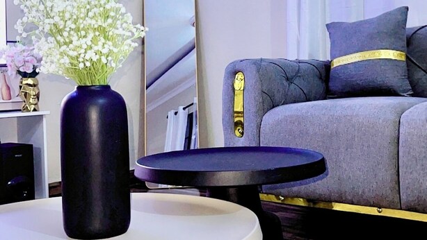 A modern living room with a round white table, a dark blue vase with white flowers, a grey sofa with yellow accents, and a large mirror on the wall. - We offer peace & serenity…wellcome to our home away from hom (Dar es Salaam)