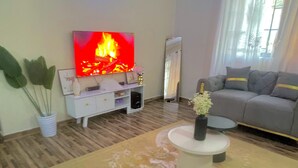 A living room with a flat-screen TV displaying a fire, a grey sofa, a white entertainment unit, and a small round coffee table with a vase. - We offer peace & serenity…wellcome to our home away from hom (Dar es Salaam)