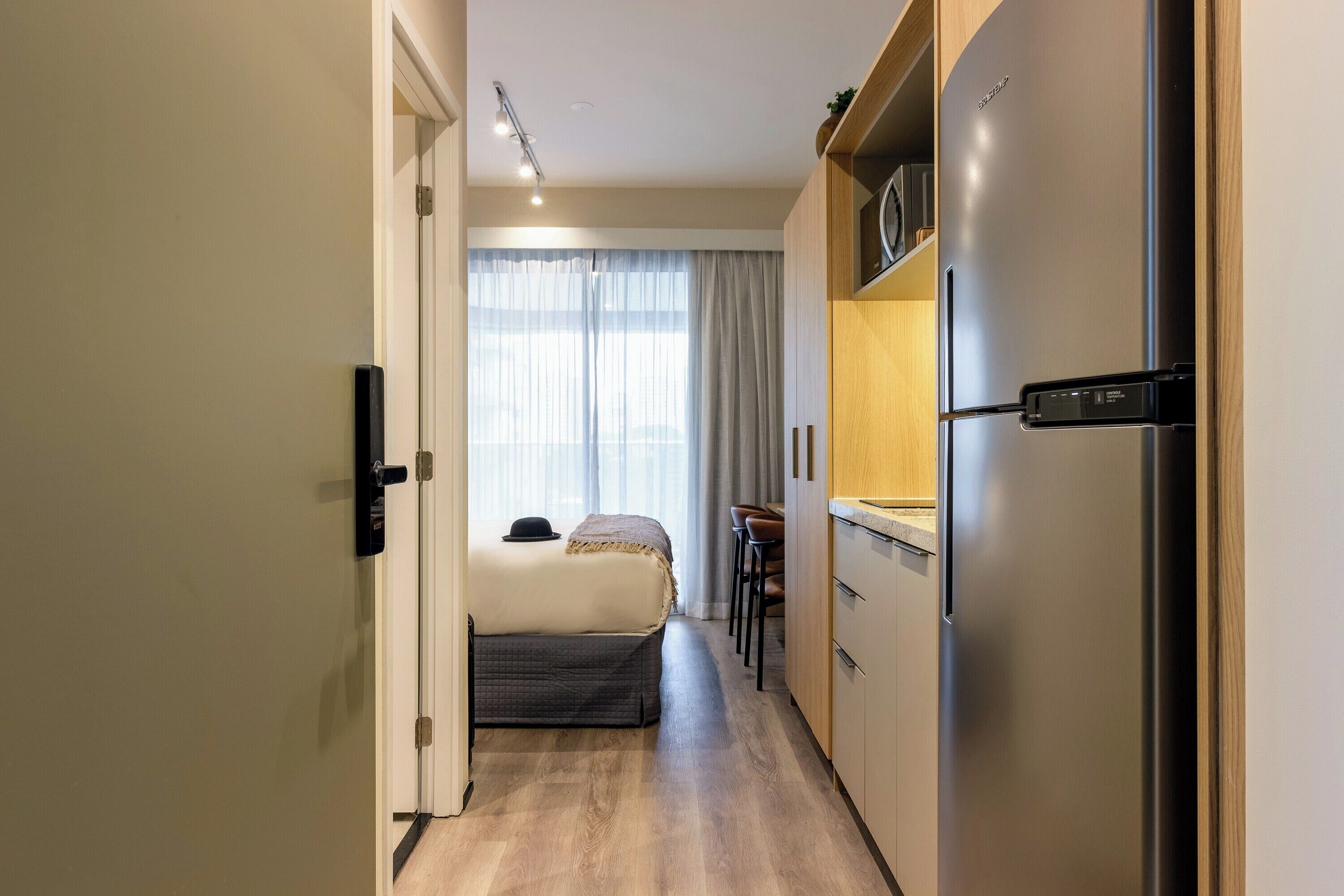 A compact hotel room with a kitchenette, a bed with a headboard, a window with curtains, and a small seating area.