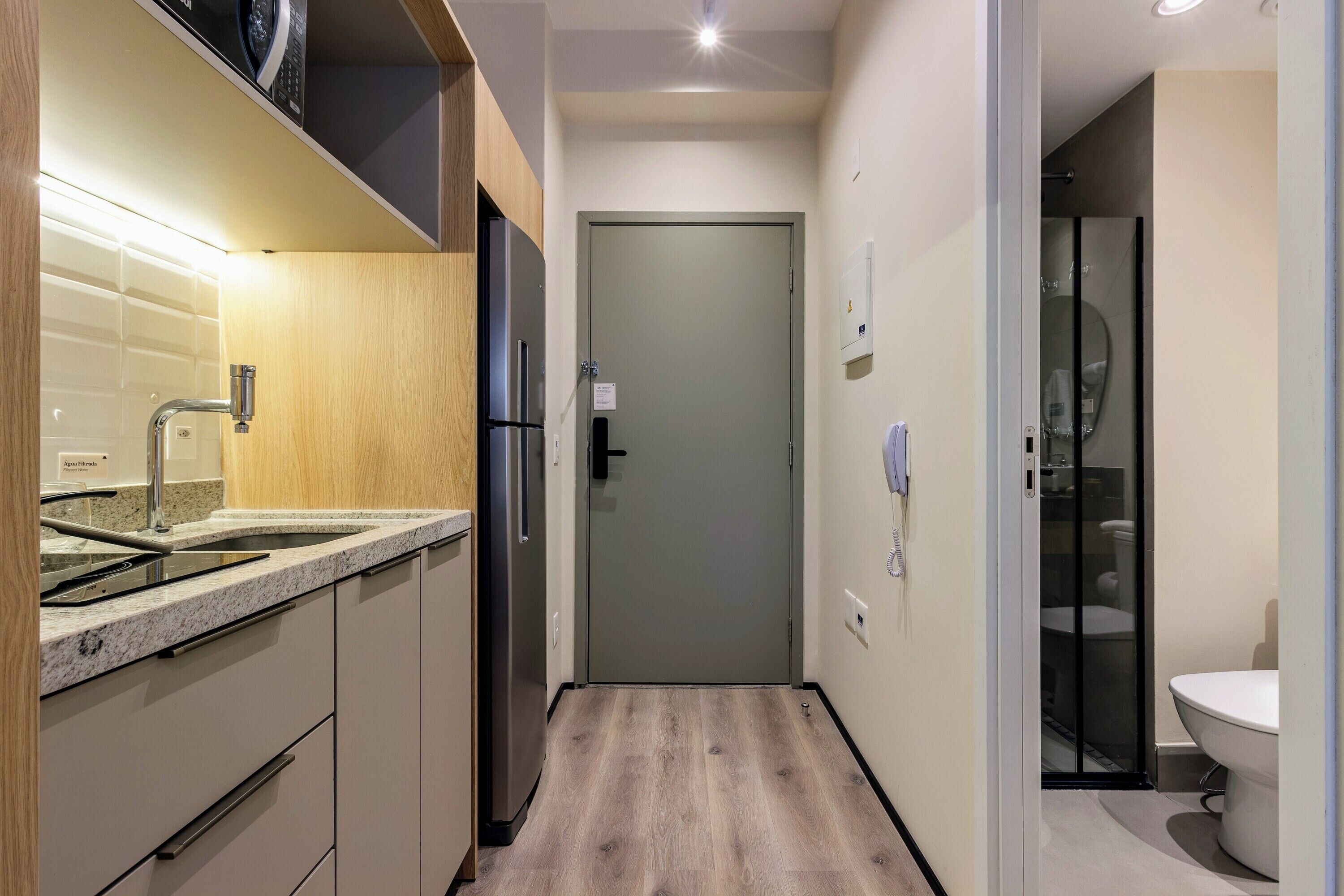 Un corridoio stretto con un angolo cottura da un lato, una porta che dà sul bagno e un design moderno caratterizzato da pareti chiare e pavimento in legno.