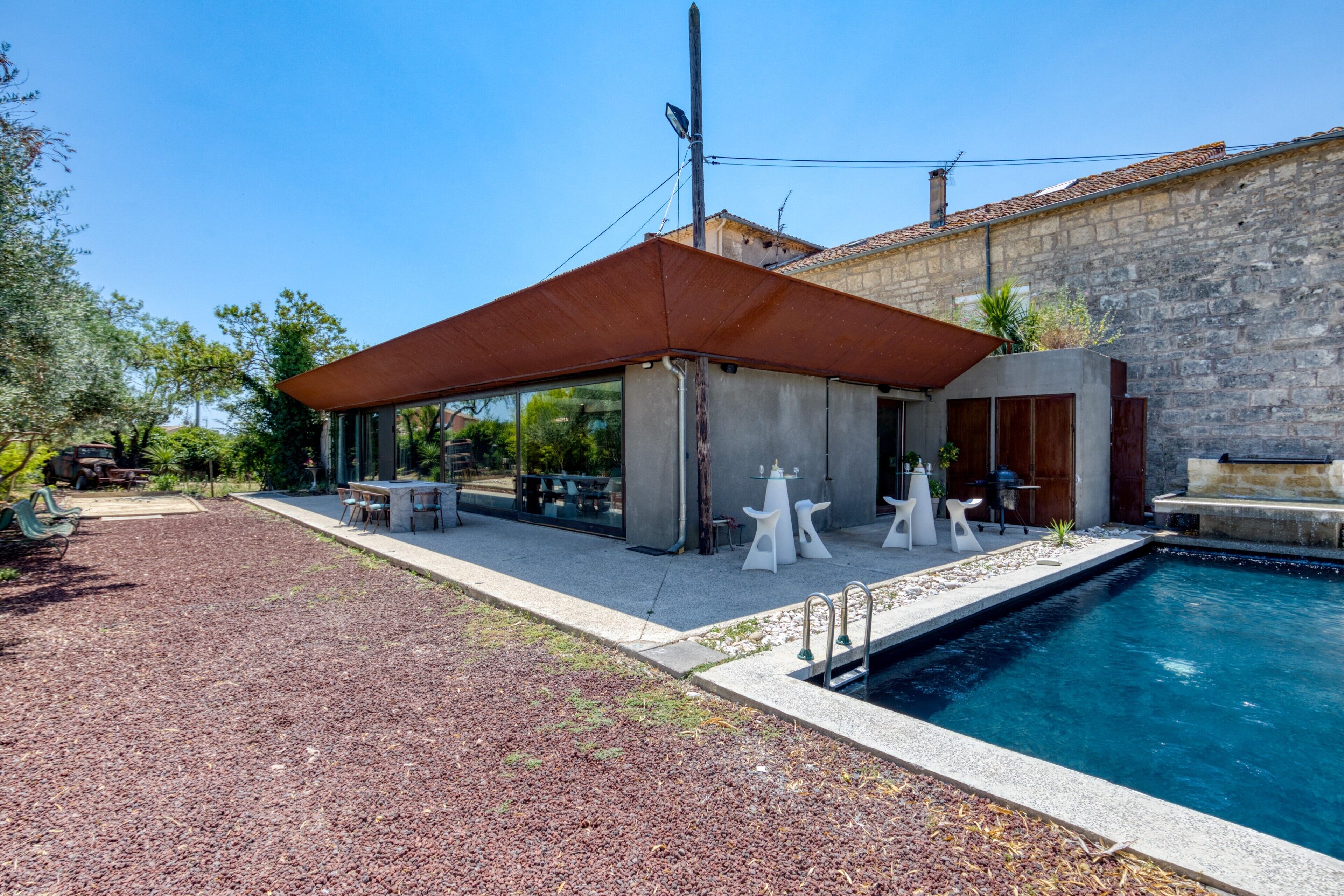 A modern outdoor pool area with a covered seating area, a stone building in the background, and a clear blue sky.