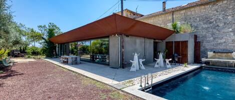 A modern outdoor pool area with a covered seating area, a stone building in the background, and a clear blue sky.