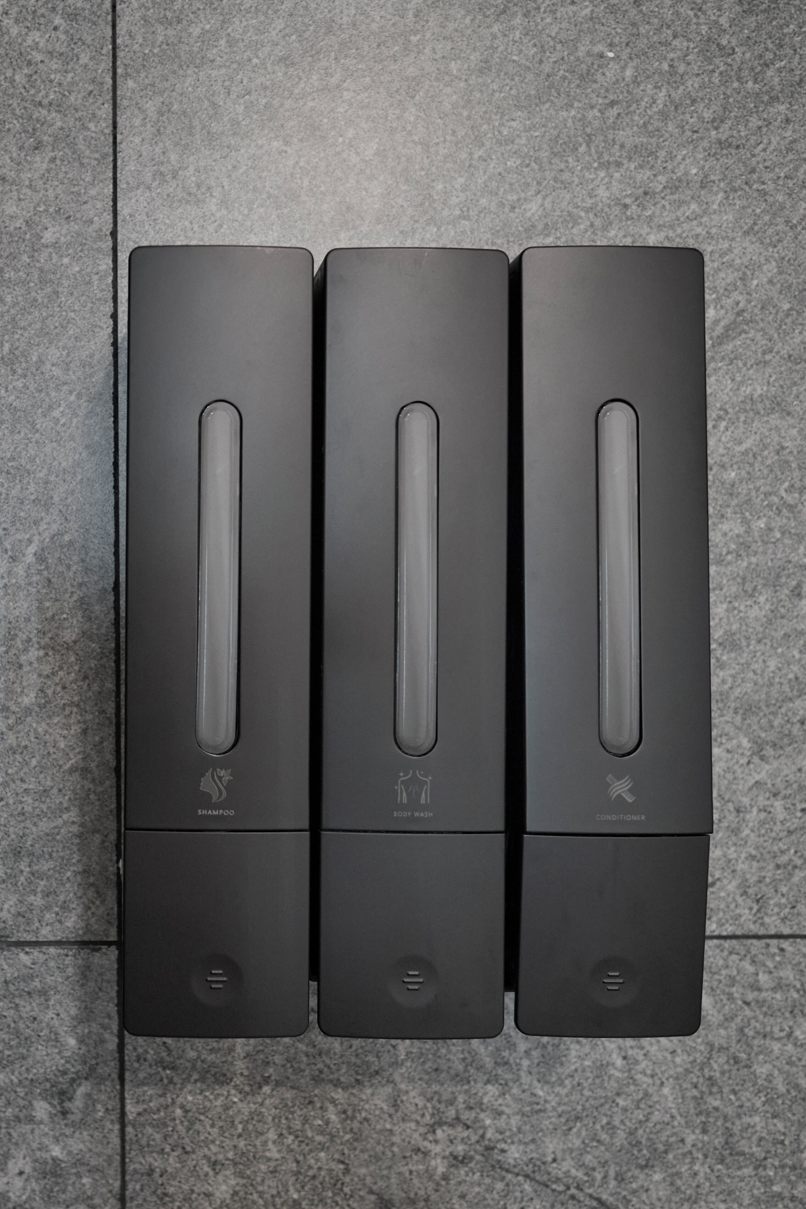 Three black, rectangular, wall-mounted dispensers labeled 'SHAMPOO,' 'BODY WASH,' and 'CONDITIONER' on a tiled floor.