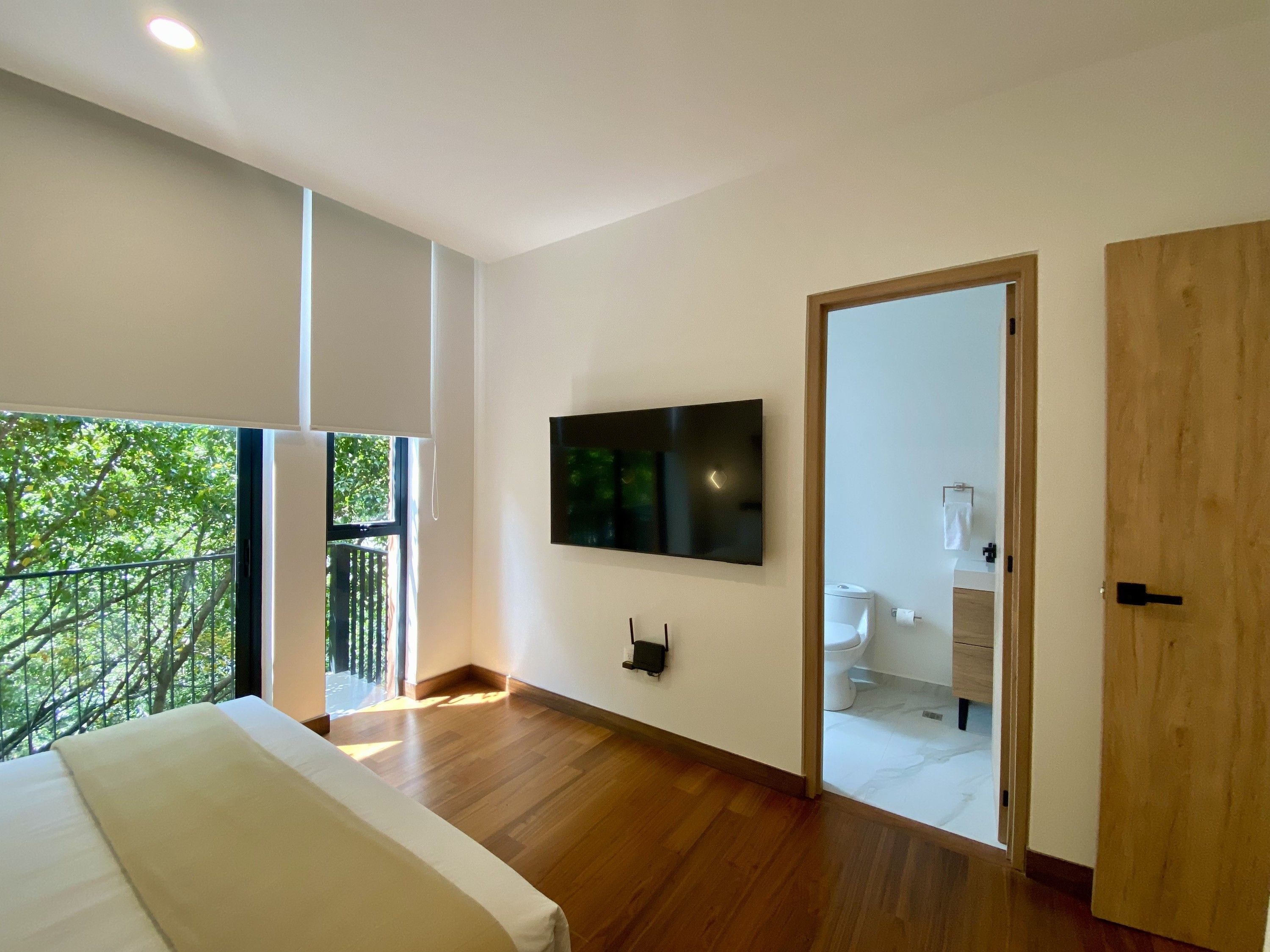 A bedroom with a large bed, a TV mounted on the wall, and a balcony with a view of trees.