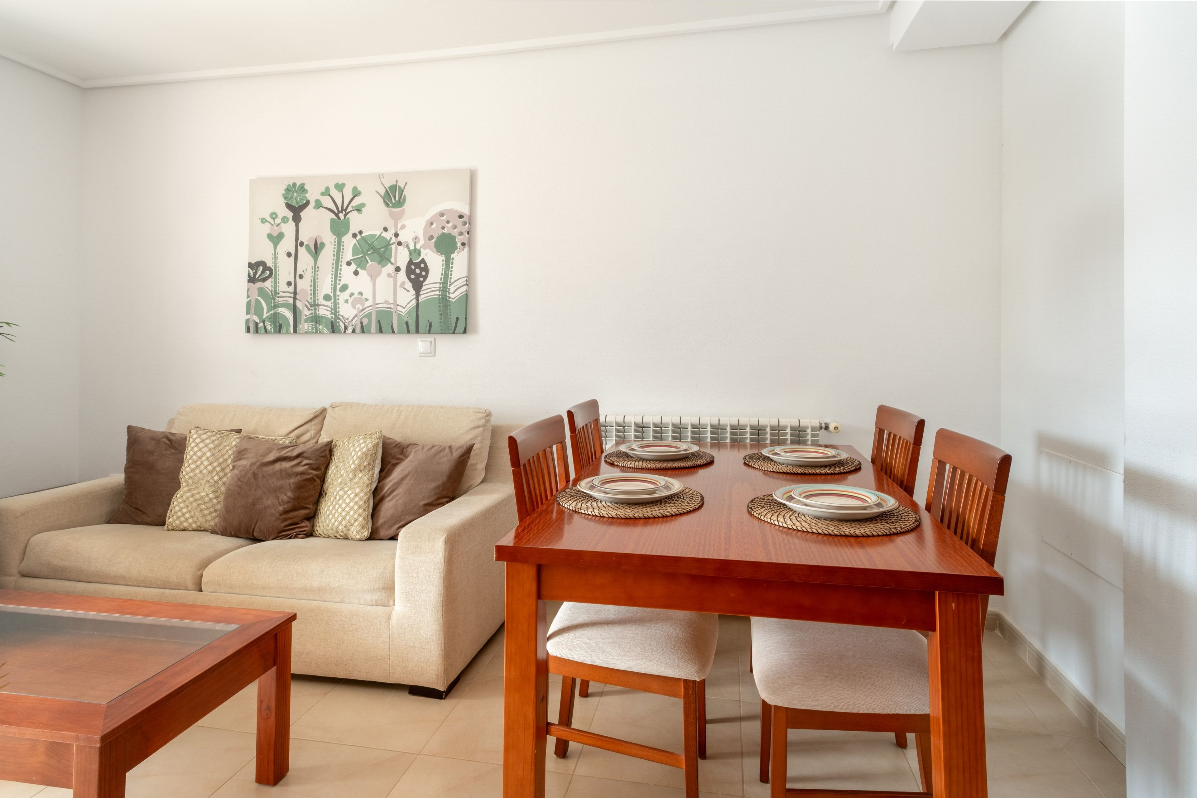 Un comedor con una mesa de madera dispuesta para cuatro, un sofá beige con cojines y un cuadro colgado en la pared.