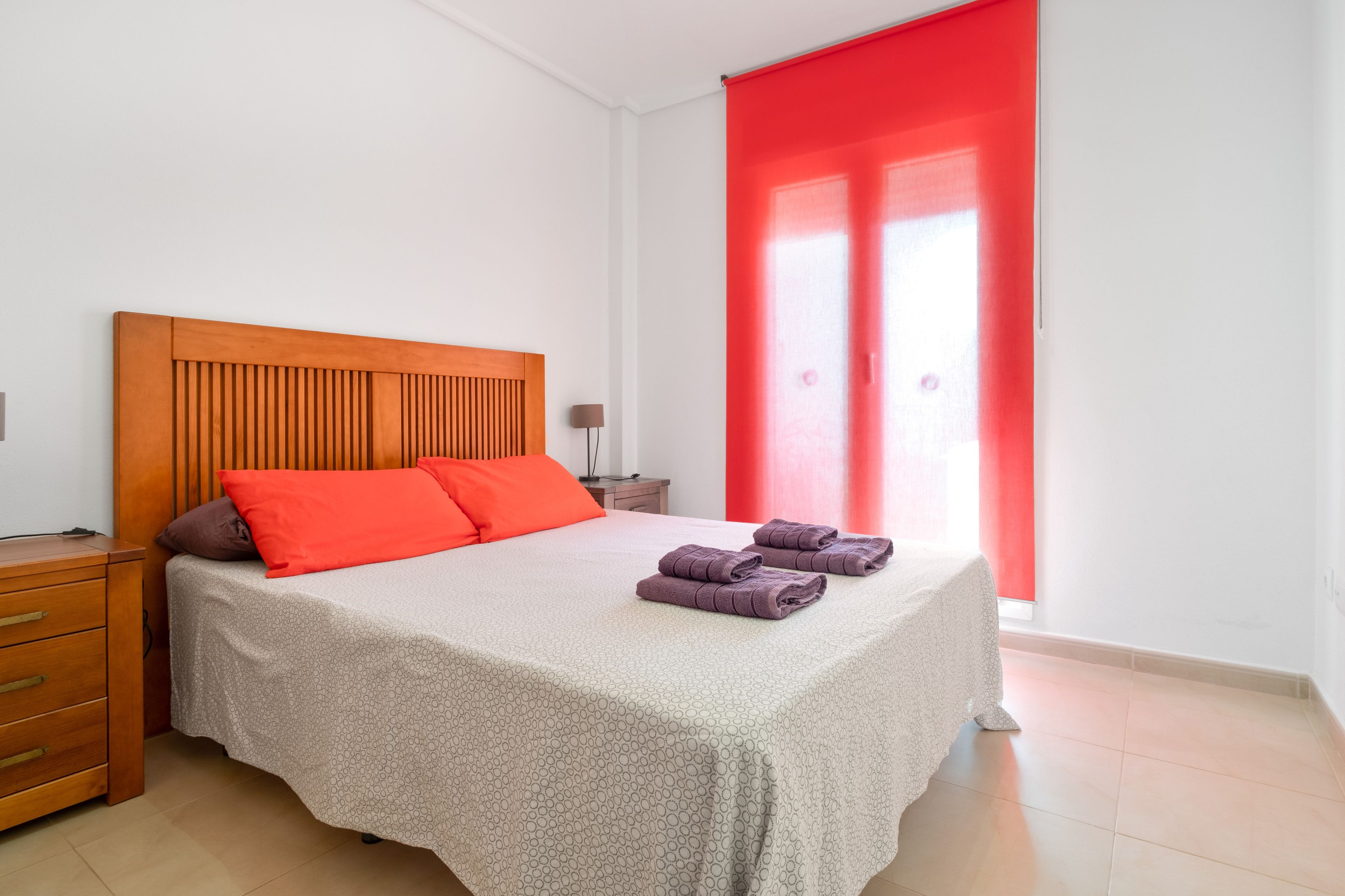 Un dormitorio con cabecera de madera, cama con ropa de cama estampada, mesita de noche, lámpara y cortina roja.