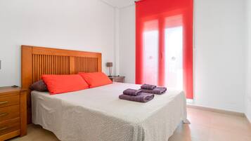 Un dormitorio con cabecera de madera, cama con ropa de cama estampada, mesita de noche, lámpara y cortina roja.