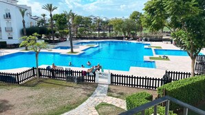 A large outdoor swimming pool with multiple lanes, surrounded by a black fence, palm trees, and a white building in the background.