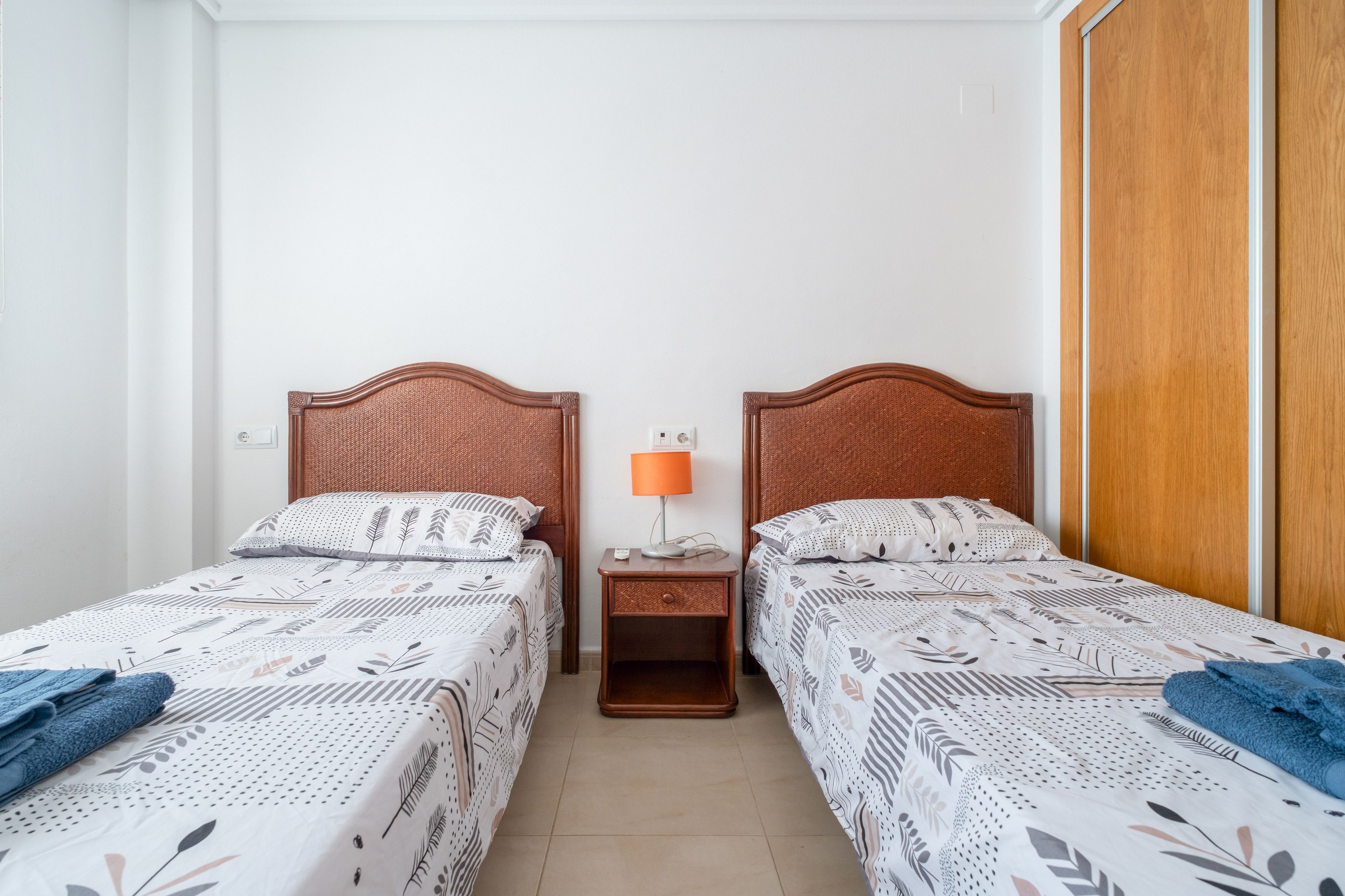 Habitación doble con dos camas individuales, una mesita de noche con lámpara y un armario de madera.