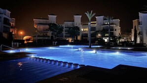 A swimming pool at night with illuminated edges, surrounded by buildings with balconies and palm trees.