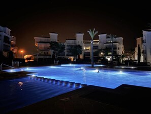 Une piscine la nuit, avec des bordures illuminées, entourée d’immeubles dotés de balcons et de palmiers.