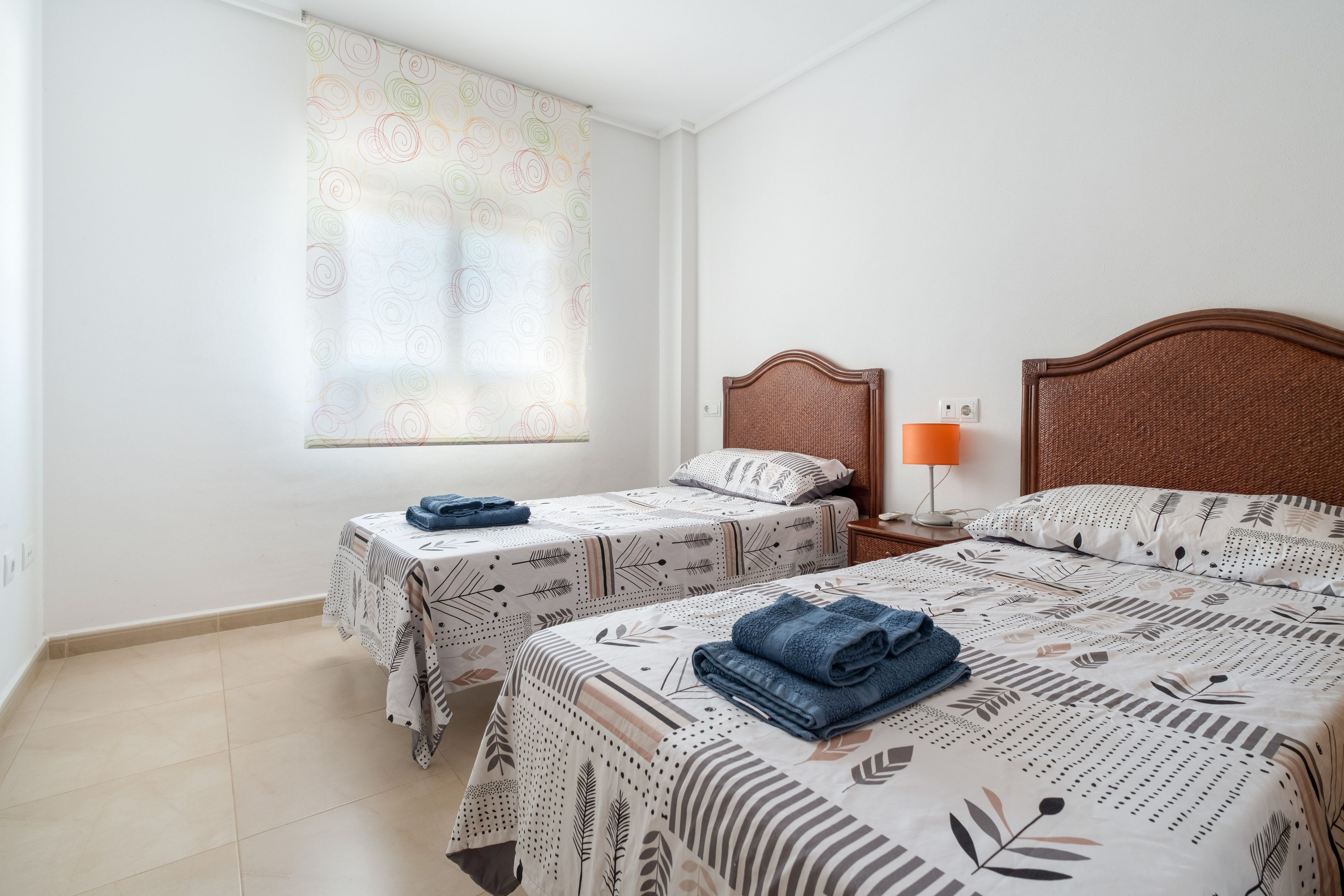 A hotel room with two beds, a window with a patterned curtain, a nightstand with a lamp, and a tiled floor.