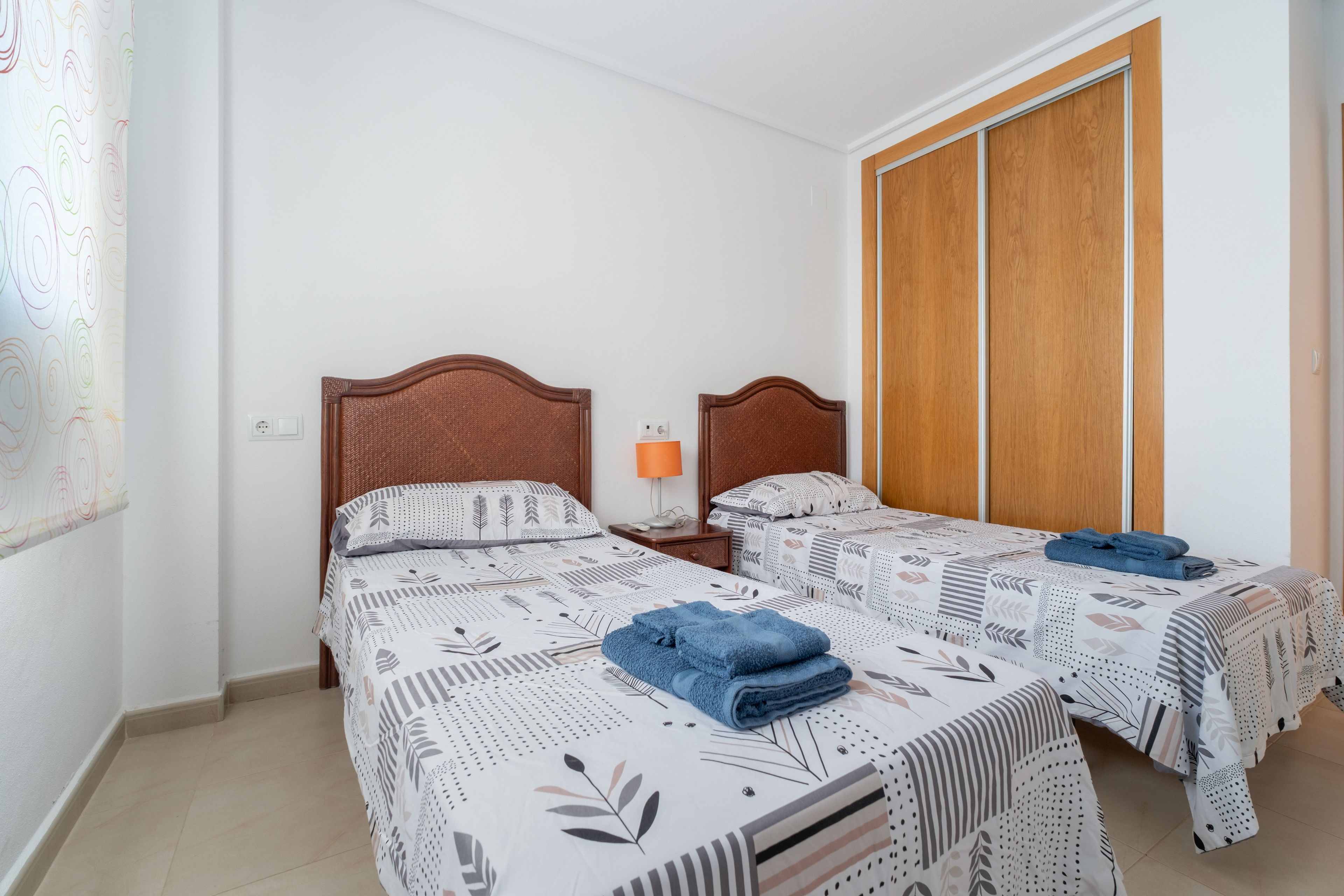 Una habitación doble con dos camas, un armario de madera, una mesita de noche con lámpara y un papel tapiz estampado.