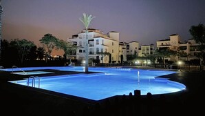 A swimming pool at night with a palm tree in the center, surrounded by buildings.