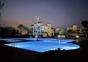 Une piscine illuminée la nuit, avec un palmier au centre, entourée d’immeubles.