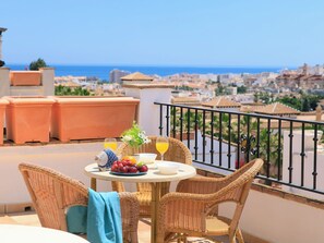 Ein Balkon mit Korbmöbeln, einem Tisch mit Obst und Getränken und Blick auf Stadt und Meer.