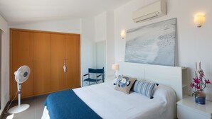 Une chambre d’hôtel comprenant un lit, un ventilateur sur pied, une armoire en bois, une chaise, un climatiseur fixé au mur et un tableau.